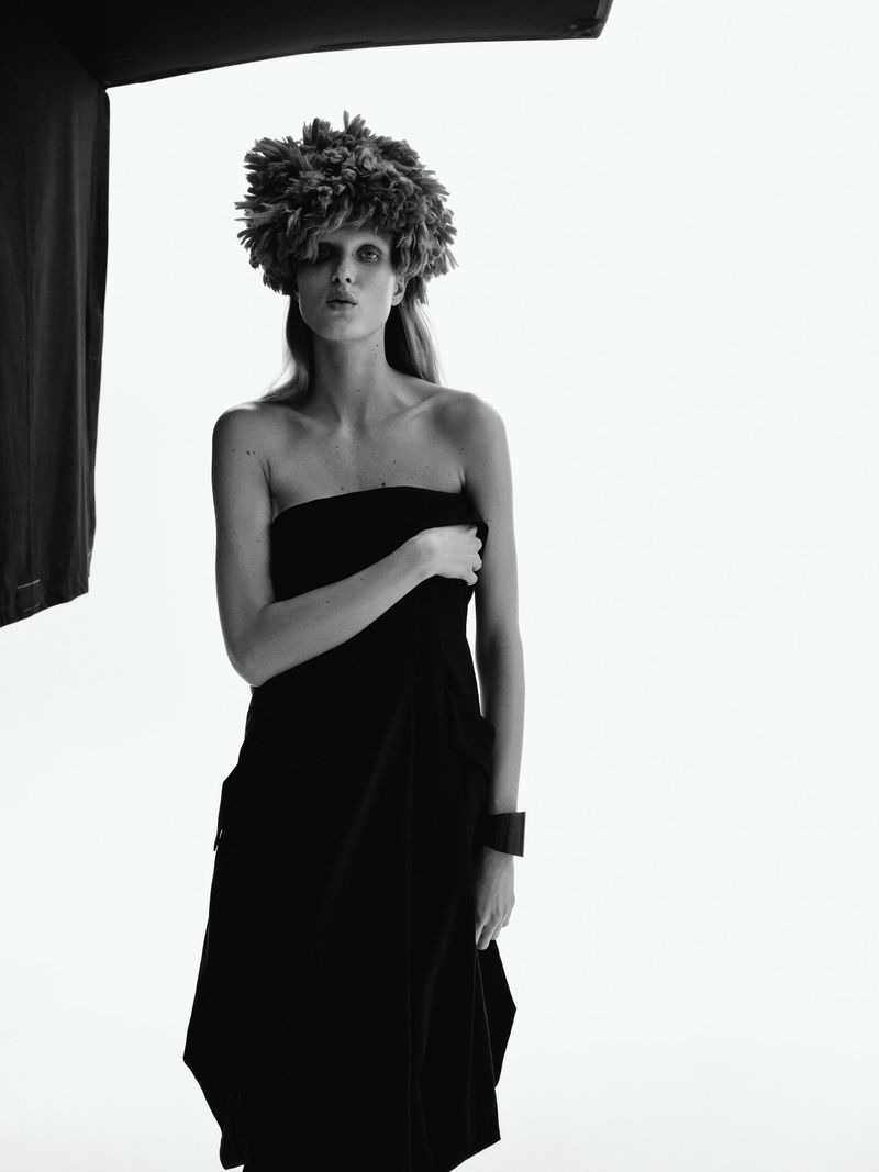 Black and white fashion portrait of a model wearing a floral headpiece and draped black fabric against a bright, soft white background.