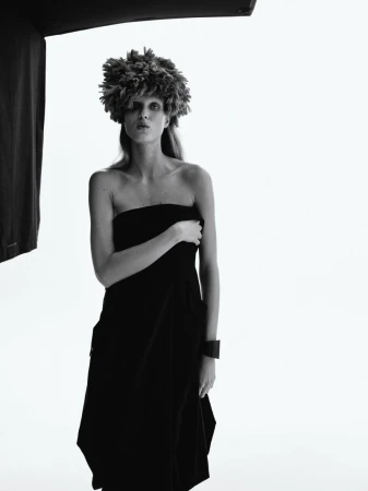 Black and white fashion portrait of a model wearing a floral headpiece and draped black fabric against a bright, soft white background.