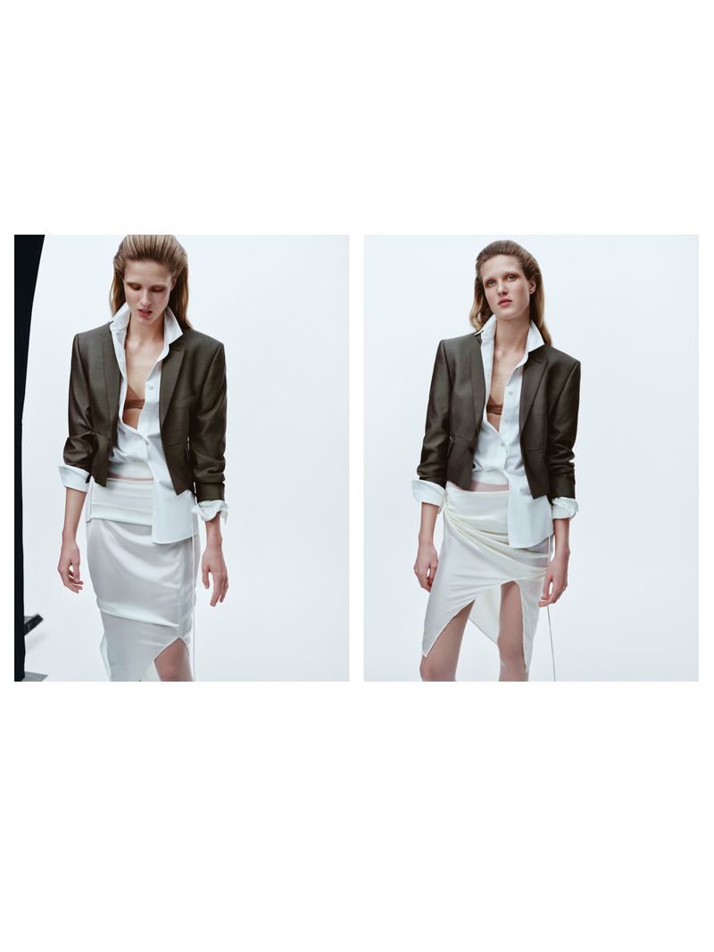 Fashion shot: Woman posing indoors in a dark cropped blazer, white button-down shirt, and a skirt with a slit, on a white background.