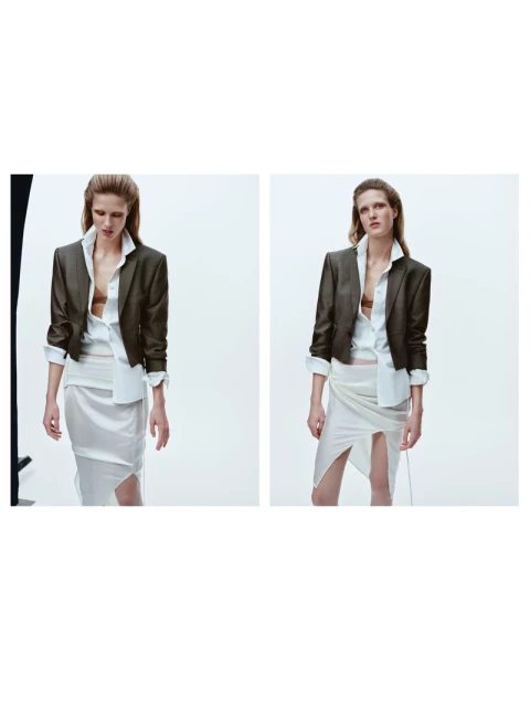 Fashion shot: Woman posing indoors in a dark cropped blazer, white button-down shirt, and a skirt with a slit, on a white background.