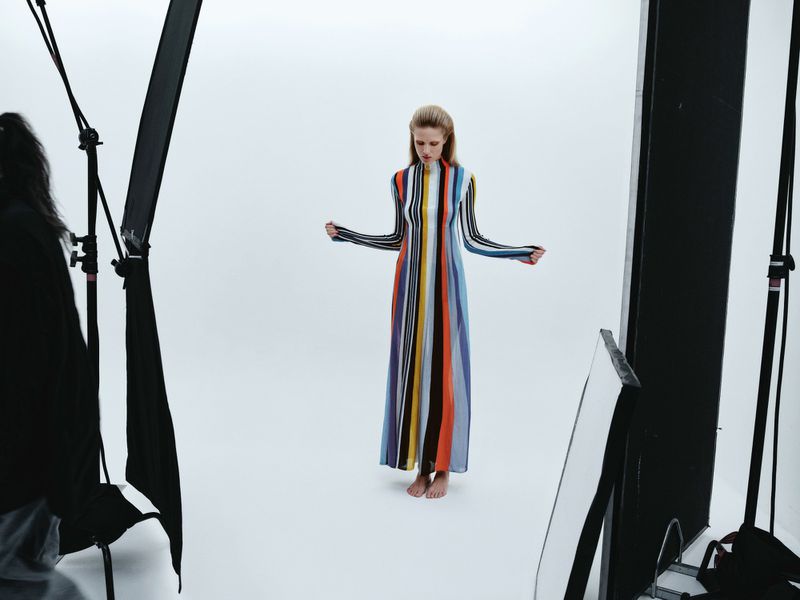 A fair-skinned adult woman with long hair poses in a floor-length striped dress in a bright photo studio with softbox lighting and black stands.