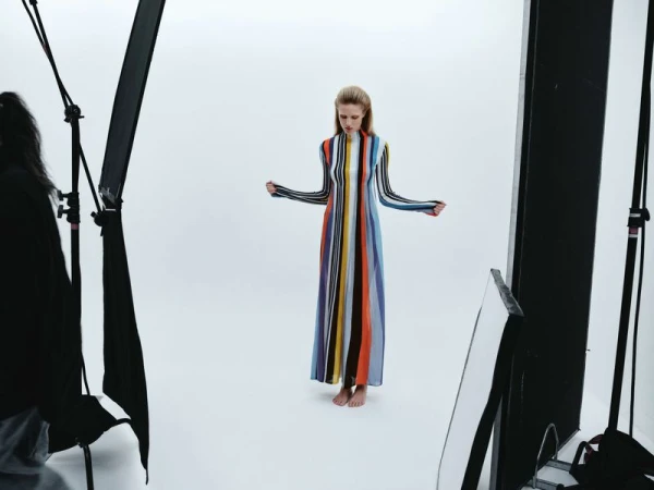 A fair-skinned adult woman with long hair poses in a floor-length striped dress in a bright photo studio with softbox lighting and black stands.