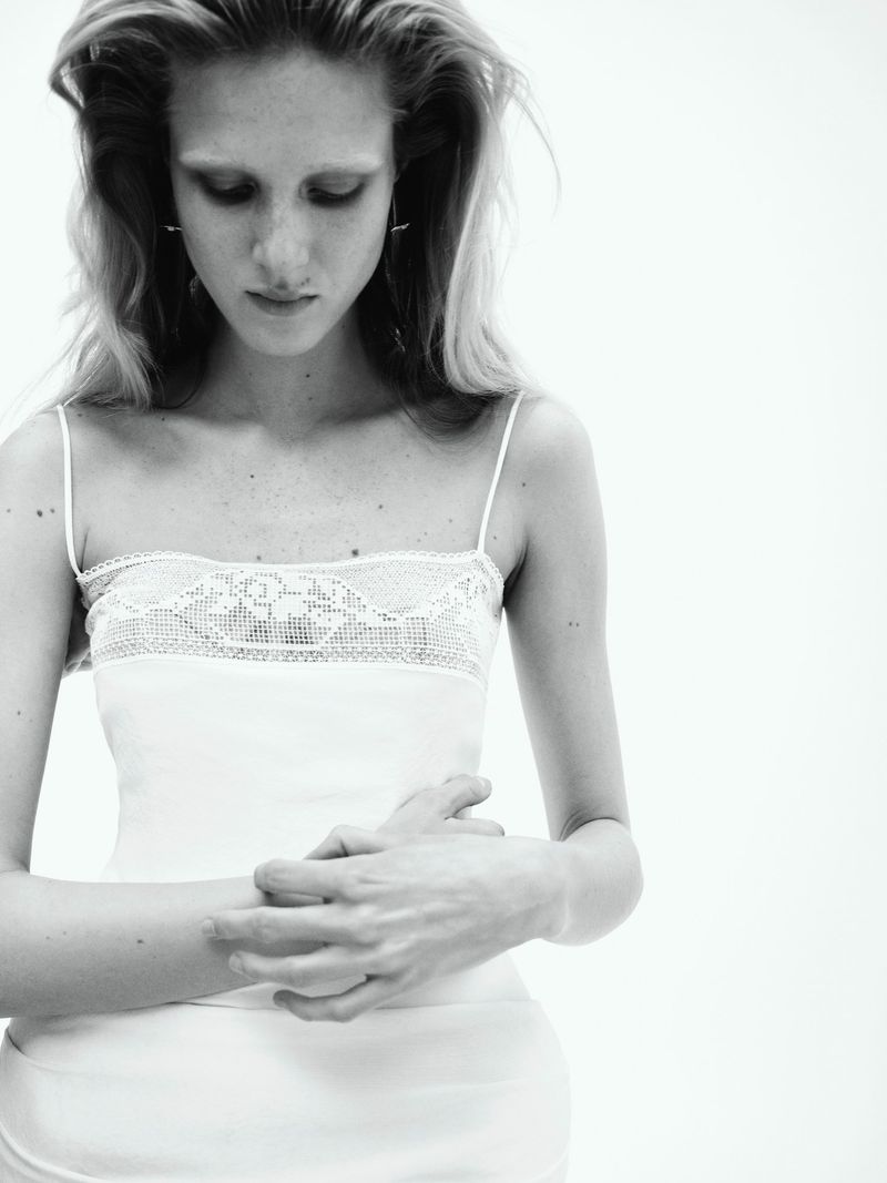 Black and white portrait of a slender, fair-skinned adult female with long wavy hair, wearing a white camisole and looking down.