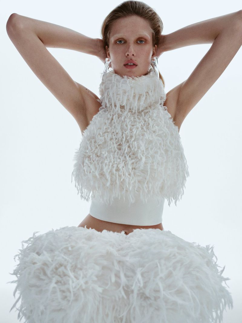 Fashion shot of a slender woman in a white, feathery two-piece outfit, standing in a bright photo studio with her arms up.