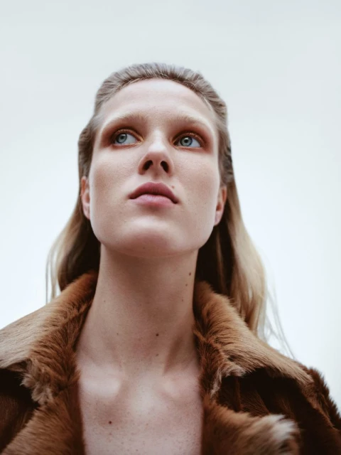 Close-up portrait of a blonde-haired woman wearing a fur collar jacket, looking up against a light background. Fashion and beauty shot.