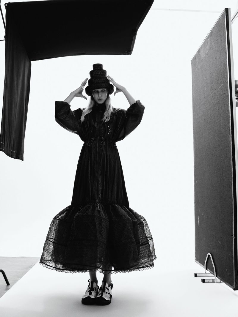 Fashion photography in black and white features a woman wearing a long black dress and a stacked hat, posing in a bright studio space.