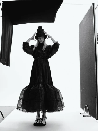 Fashion photography in black and white features a woman wearing a long black dress and a stacked hat, posing in a bright studio space.