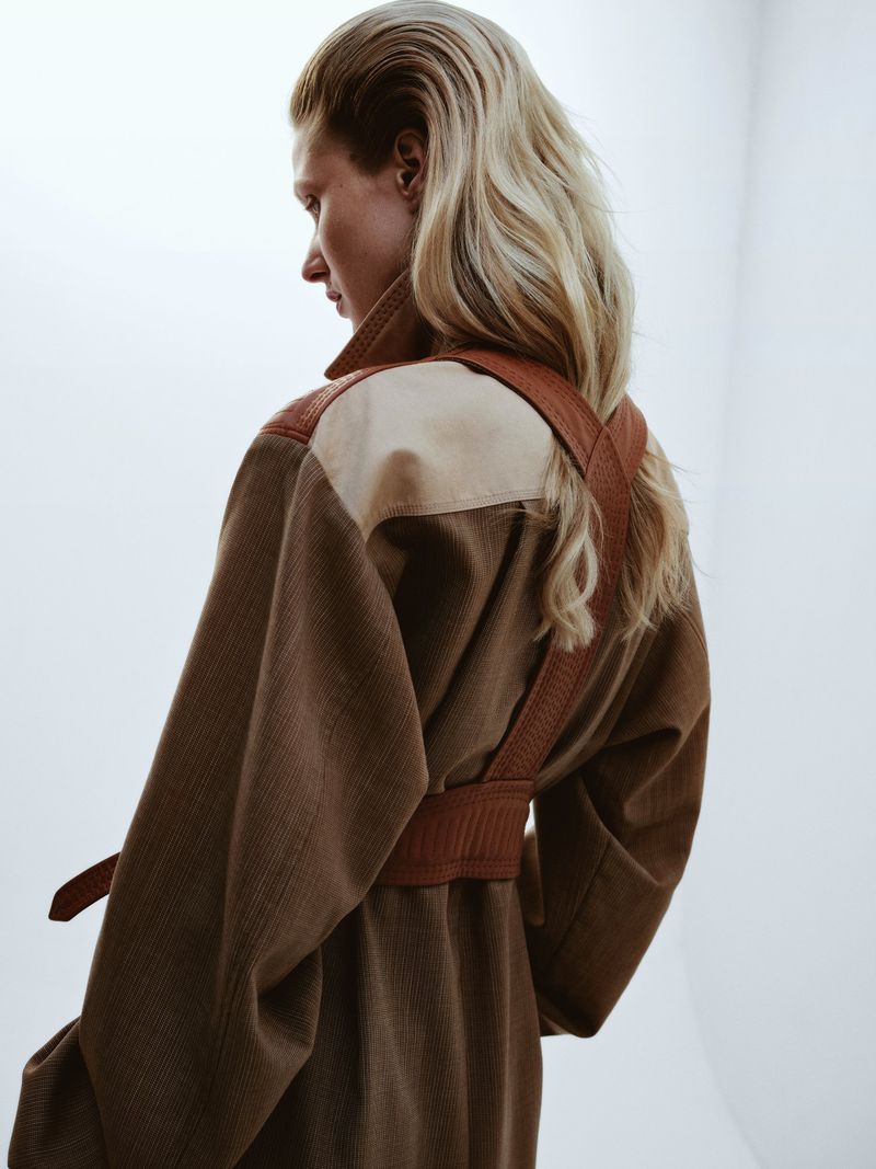 Back view of a blonde adult female wearing a brown overcoat with matching belt in a studio setting against a white backdrop.