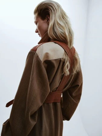 Back view of a blonde adult female wearing a brown overcoat with matching belt in a studio setting against a white backdrop.