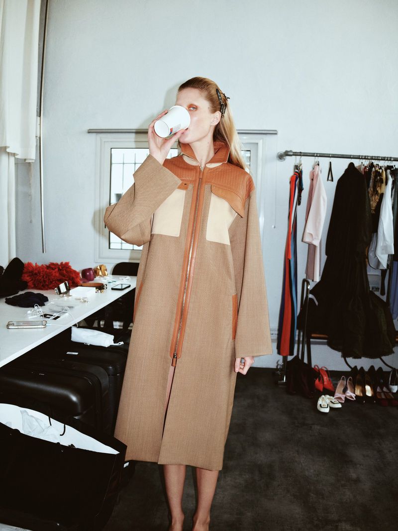 Woman in a tan coat drinks from a disposable cup in a cluttered dressing room with clothes rack, luggage, table, and shoes.
