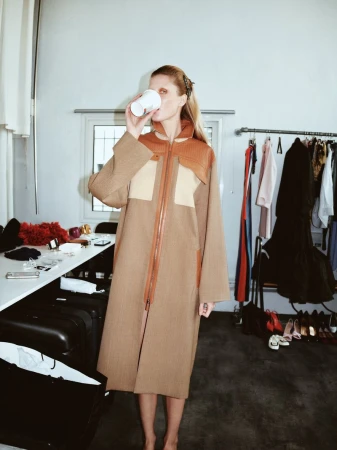 Woman in a tan coat drinks from a disposable cup in a cluttered dressing room with clothes rack, luggage, table, and shoes.