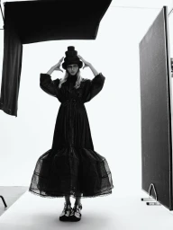 Model wearing a structured dark dress and tiered hat in a studio setting with lighting flags
