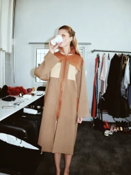 Model Vakare Steputyte drinks from a paper cup backstage at Costume Denmark wearing a long tan coat