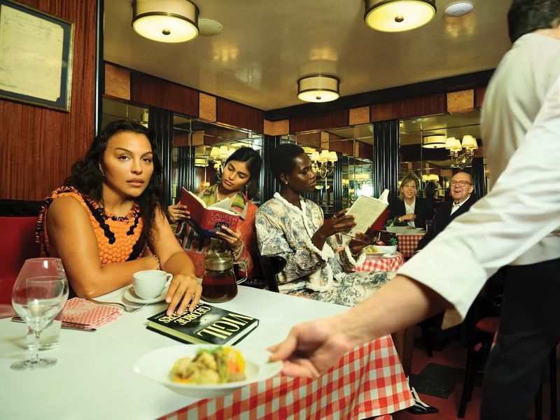 Several people are in an indoor restaurant setting with tables, chairs, and a waiter serving food in a warmly lit environment.