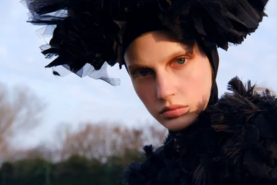 Model wearing a large black ruffled headpiece and textured dark garment outdoors against an open sky background.