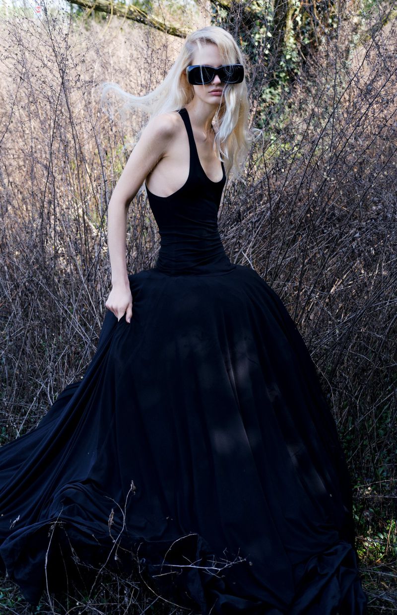 A stylish adult female with long blond hair wearing a black dress and sunglasses, standing outdoors among tall dry grasses and bare trees.
