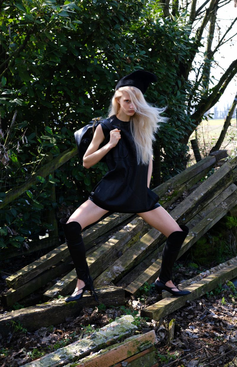 A slender, fair-skinned woman in a black dress poses dynamically on fallen wooden planks outdoors against a backdrop of trees and vegetation.