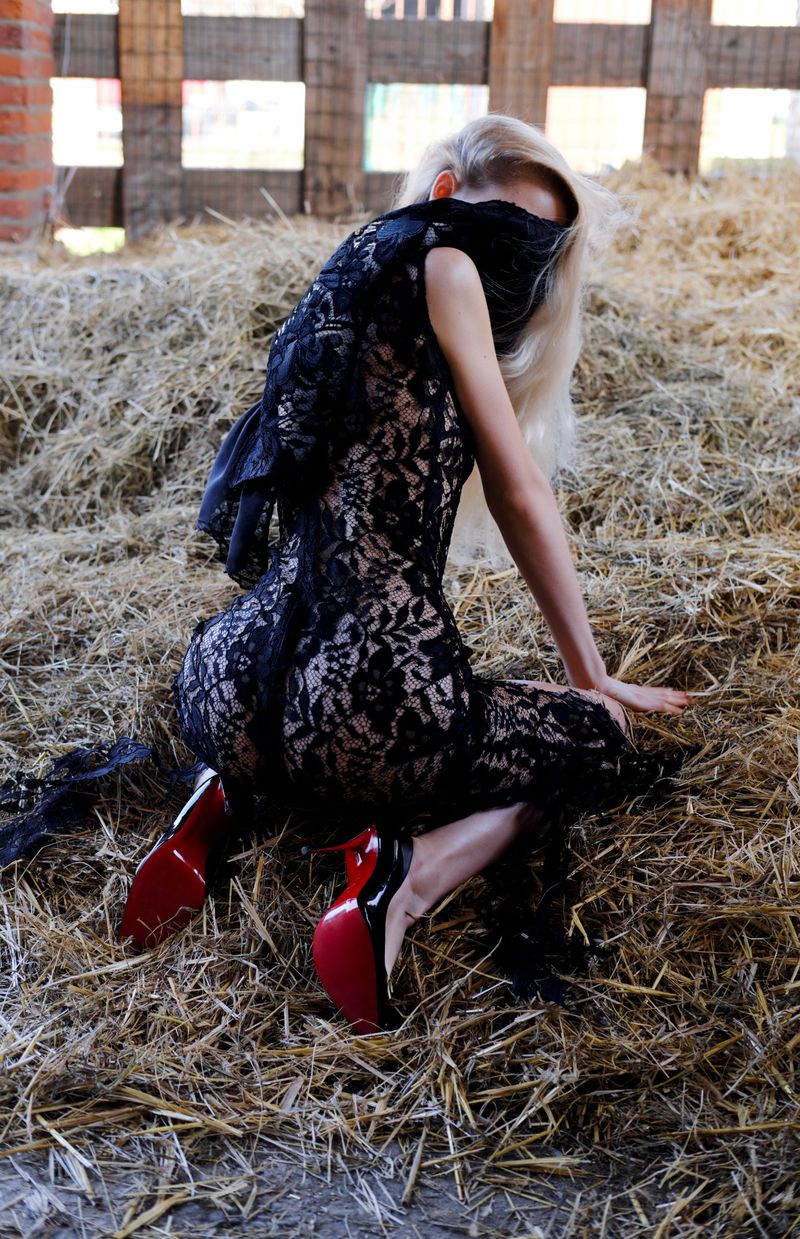 Blonde woman in black lace dress, wearing high heels, kneeling on hay with a black cloth covering her eyes in a barn.