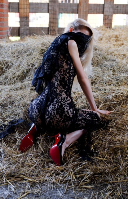 Blonde woman in black lace dress, wearing high heels, kneeling on hay with a black cloth covering her eyes in a barn.