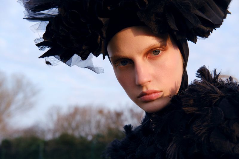 Close-up portrait of a woman with fair skin wearing a black headpiece and feathered collar, set against a blurred outdoor backdrop.