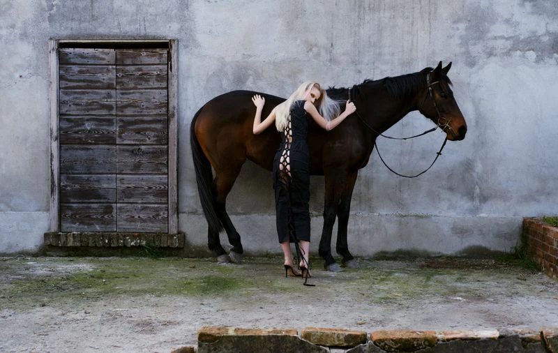 A woman in a black dress stands next to a brown horse against a weathered concrete wall in an outdoor environment with an old door.