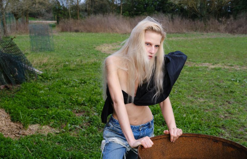 A woman with blond hair poses in a black top and ripped jeans in an outdoor setting near a rusted barrel, with a green grassy background.
