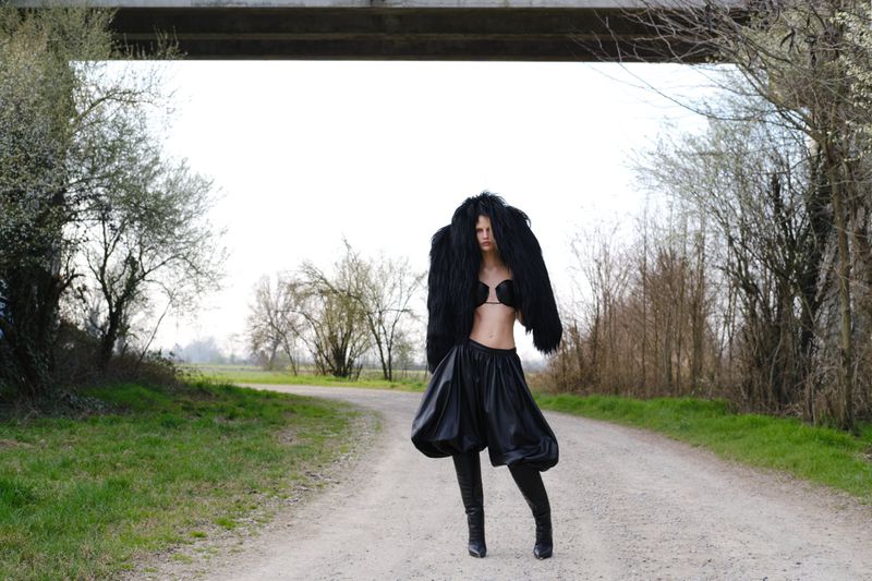 A slender woman in a black bra, fur coat, and balloon shorts strikes a pose on a gravel path near green fields and an overpass.