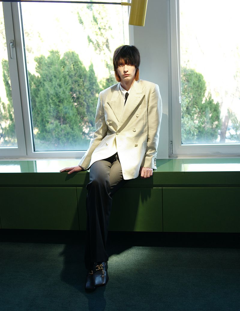 A young adult sits against a window wearing a white patterned blazer, a tie, and black pants. The lighting is soft and diffused.