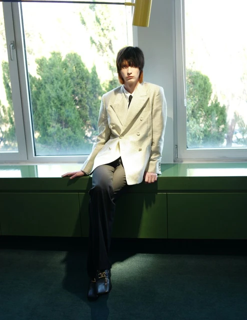 A young adult sits against a window wearing a white patterned blazer, a tie, and black pants. The lighting is soft and diffused.