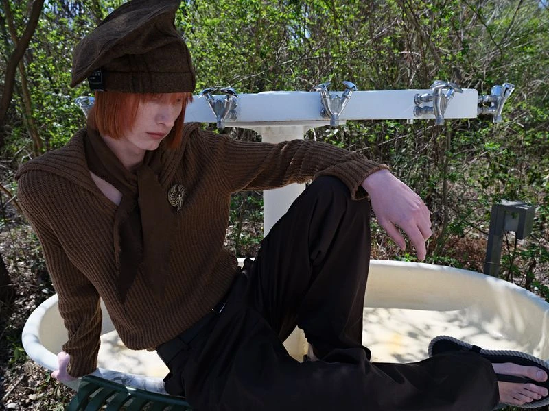 A young adult with red hair and brown clothing is sitting on an old bathtub with trees in the background on a sunny day outdoors.