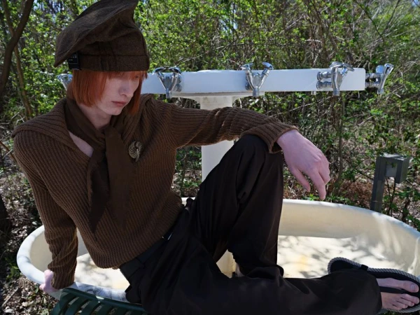 A young adult with red hair and brown clothing is sitting on an old bathtub with trees in the background on a sunny day outdoors.