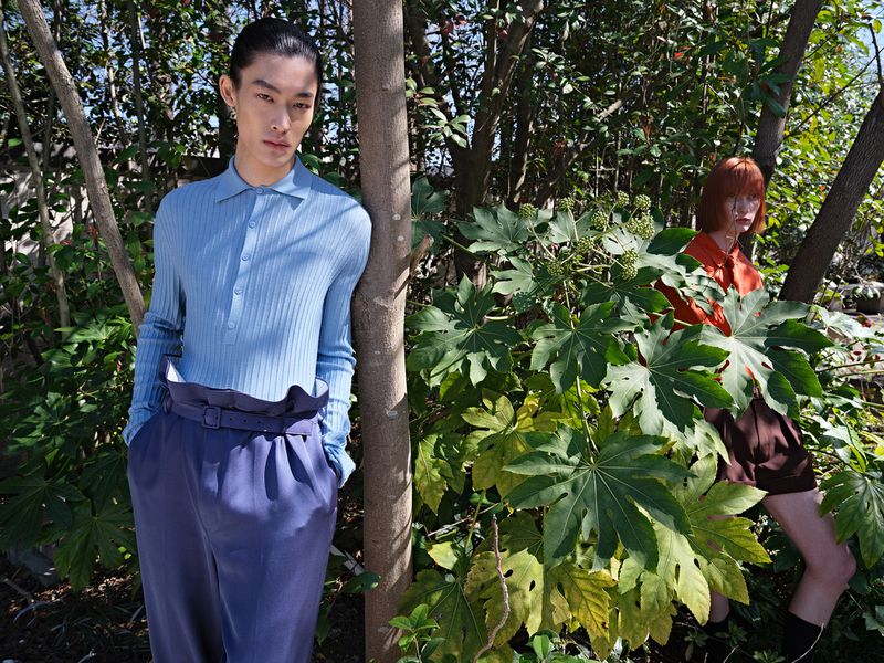 Two fashion models pose among trees and lush green plants. One wears blue ribbed shirt and pants, while the other wears orange top and skirt.
