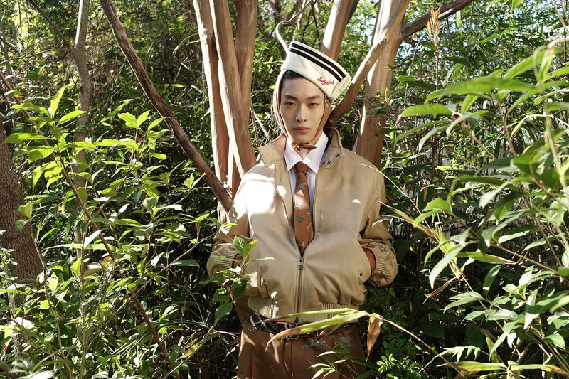 Adult male in tan jacket and sailor hat posing in a dense forest with green foliage and trees in the background, eye-level angle.