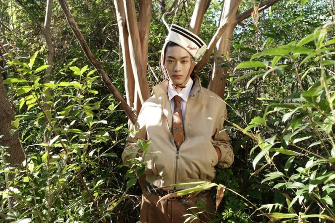 Adult male in tan jacket and sailor hat posing in a dense forest with green foliage and trees in the background, eye-level angle.