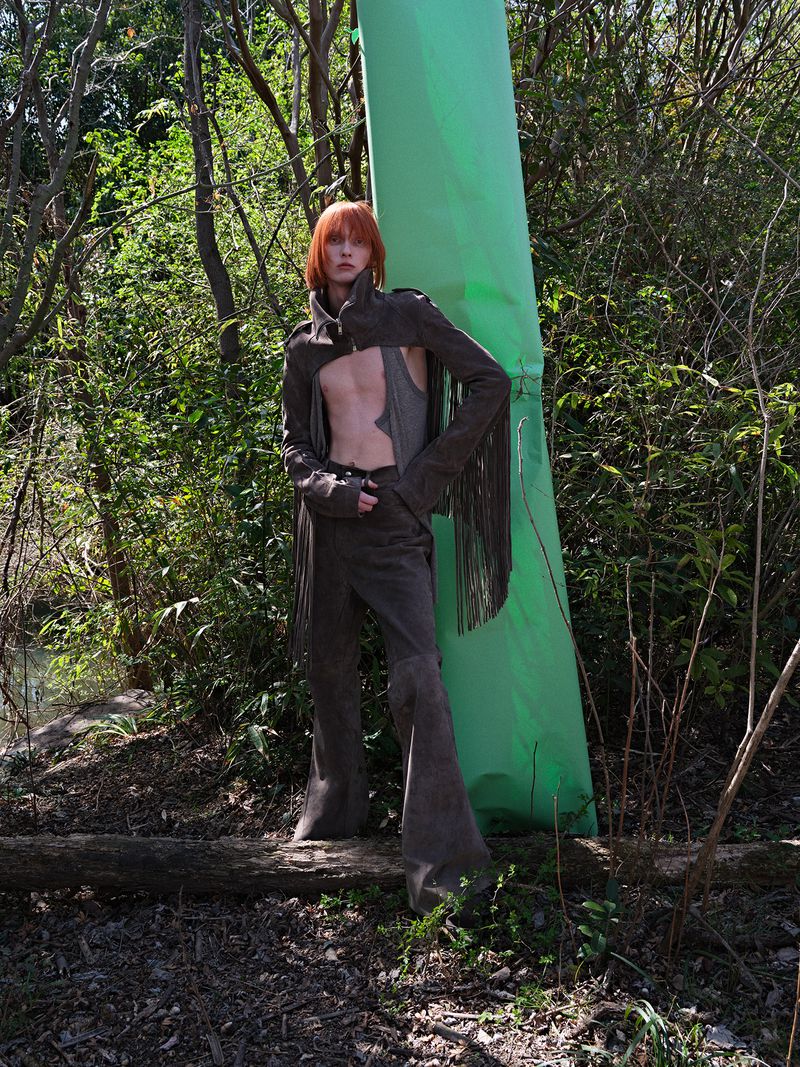 Fashion shot of a red-haired male model wearing a brown suede outfit with fringe, standing in a wooded, outdoor environment with bright sunlight.