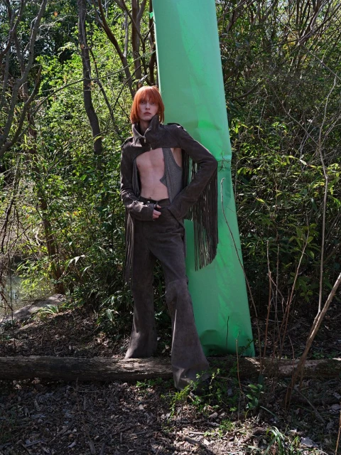 Fashion shot of a red-haired male model wearing a brown suede outfit with fringe, standing in a wooded, outdoor environment with bright sunlight.