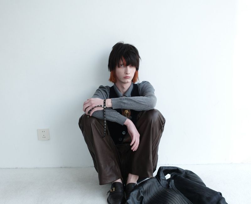 A young adult sits cross-legged against a white wall in a gray top and brown pants, complemented by black loafers and jacket.