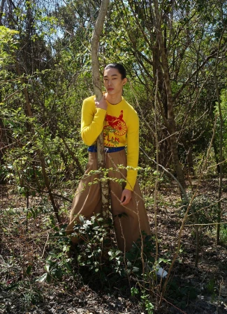 Young man in yellow long sleeve shirt poses near a thin tree in a sunny forest. He wears tan pants with blue accents and the forest surrounds him.