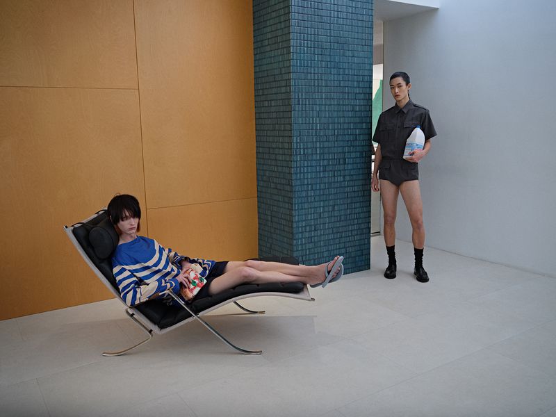 An interior shot with a woman lounging on a black chaise lounge and a man standing, holding a milk carton near a blue brick column.