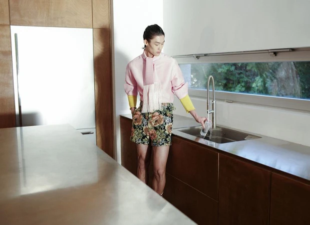 A person in a pink shirt and floral shorts stands at a kitchen sink, pouring water into a glass, with natural light filtering in.