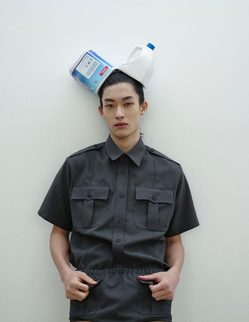 Young man poses indoors with grey button-down shirt and white plastic container on head, set against a plain white wall backdrop.