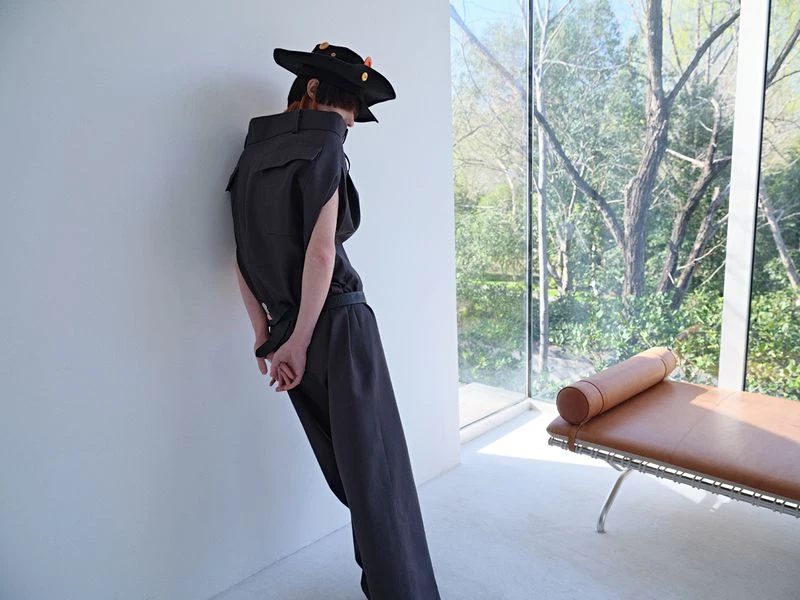 Model wearing a dark jumpsuit and hat poses against a white wall in front of a window showing trees, inside a minimalist setting.