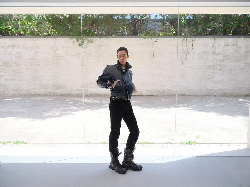 Asian person posing in stylish denim jacket, black pants, and boots indoors, illuminated by daylight in minimalist setting.