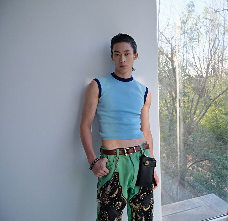 Young adult male poses in a light blue shirt and decorative pants indoors with natural light, showcasing fashion against a window background.