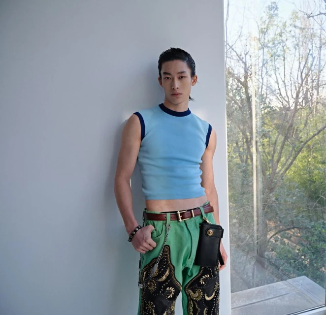 Young adult male poses in a light blue shirt and decorative pants indoors with natural light, showcasing fashion against a window background.