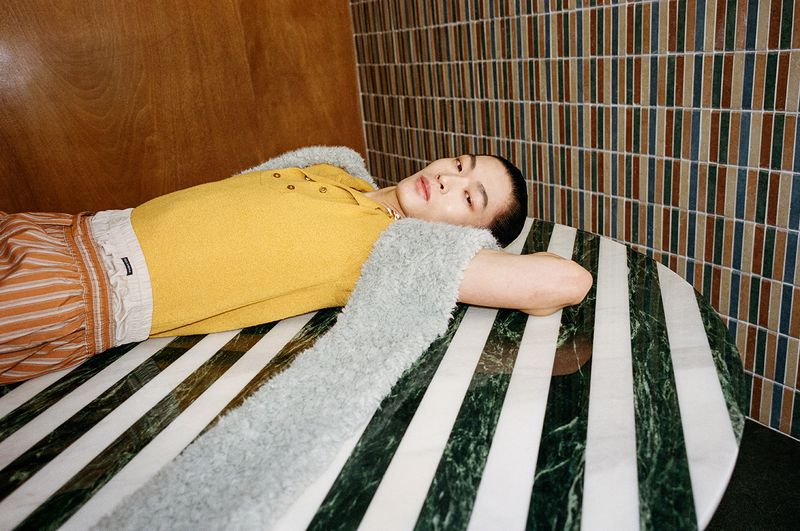 A stylish Asian male model lies on a striped table in a yellow shirt and striped pants, against a brown and tiled wall.