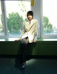 A young male model seated on a green ledge wearing a patterned white blazer and dark trousers against windows.