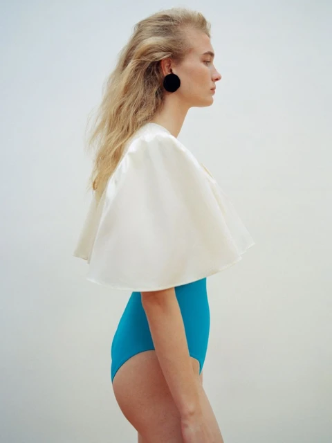 Side profile studio shot of a blond woman with a white cape and blue bathing suit against a white background in bright lighting.