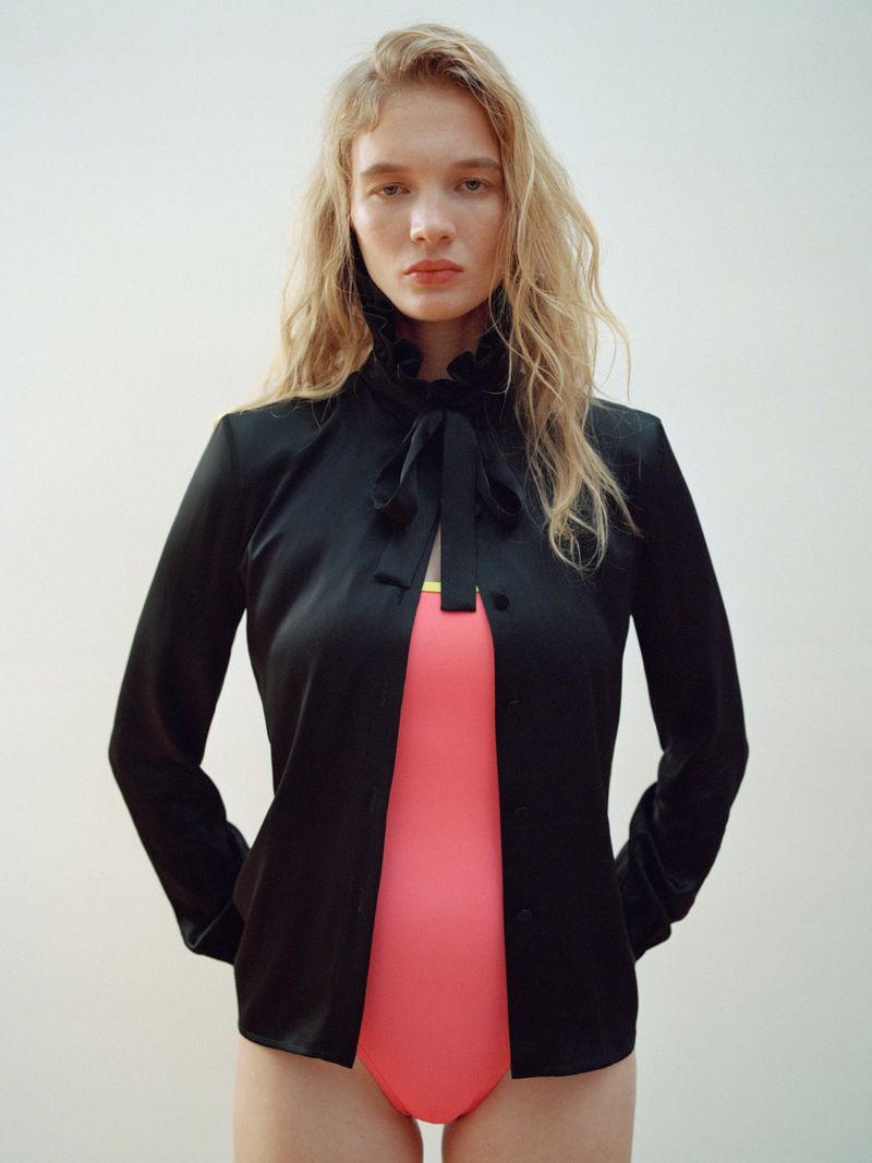 Woman in a black jacket and coral leotard stands indoors against a plain white wall, showcasing fashion and style with soft, diffused lighting.