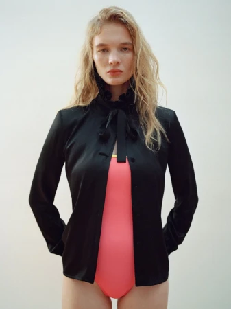 Woman in a black jacket and coral leotard stands indoors against a plain white wall, showcasing fashion and style with soft, diffused lighting.